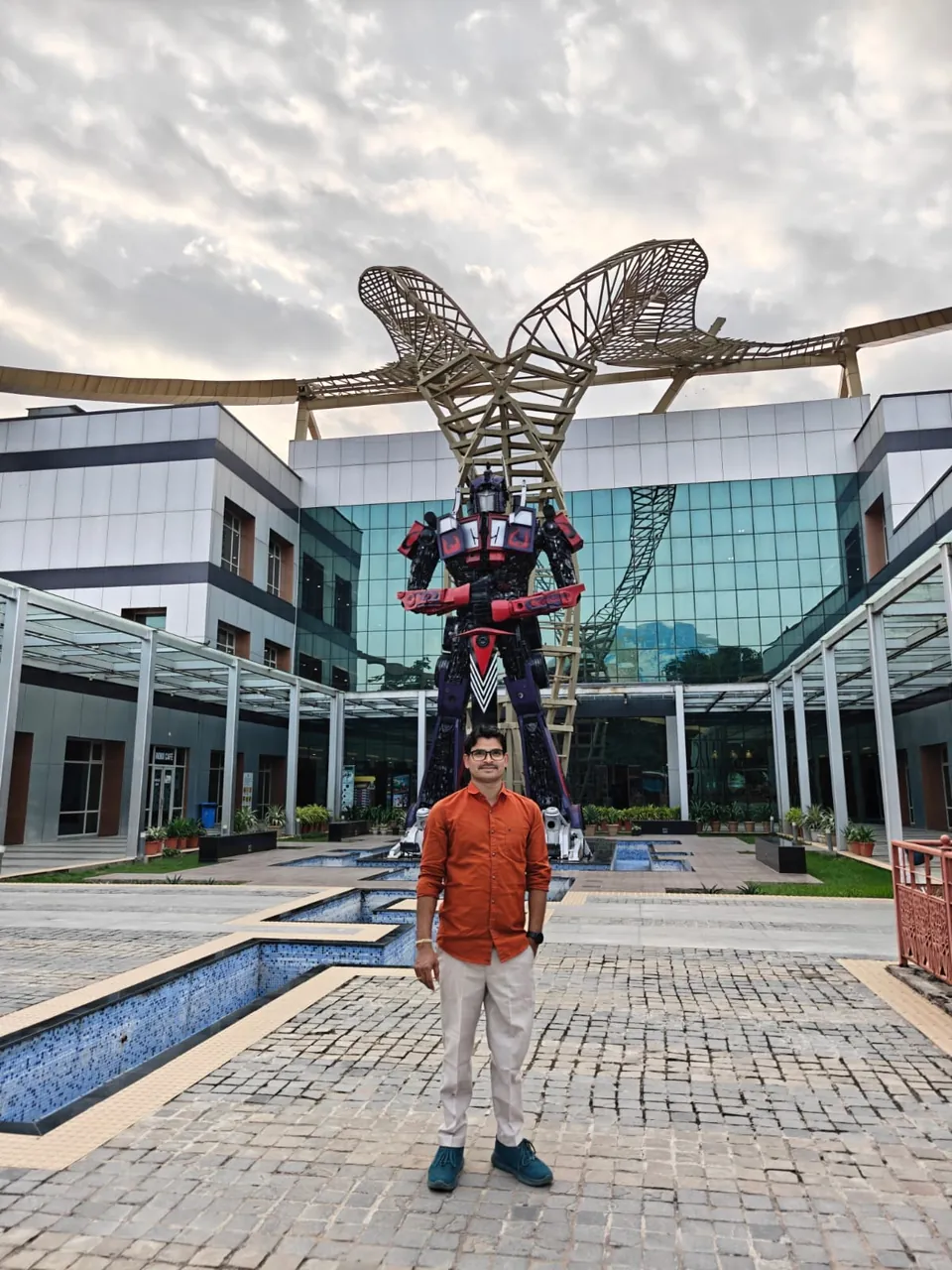 Krishna Kummari in an orange shirt standing in a paved courtyard in front of a large robot sculpture and a glass office building.