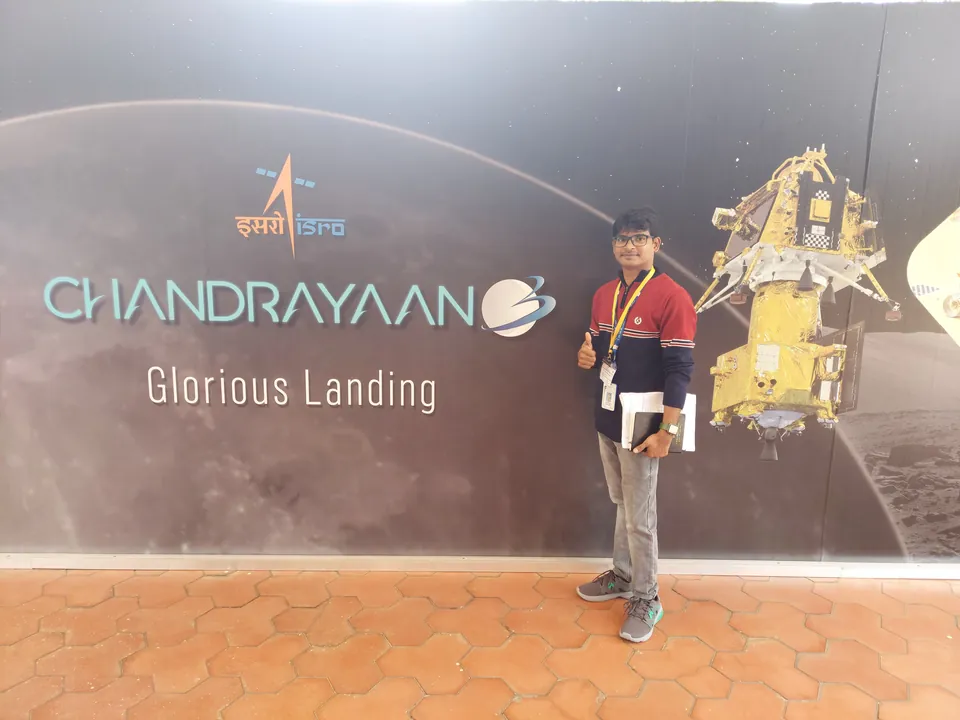 Krishna Kummari standing in front of a dark wall printed with the ISRO logo, the word Chandrayaan and the words Glorious Landing.
