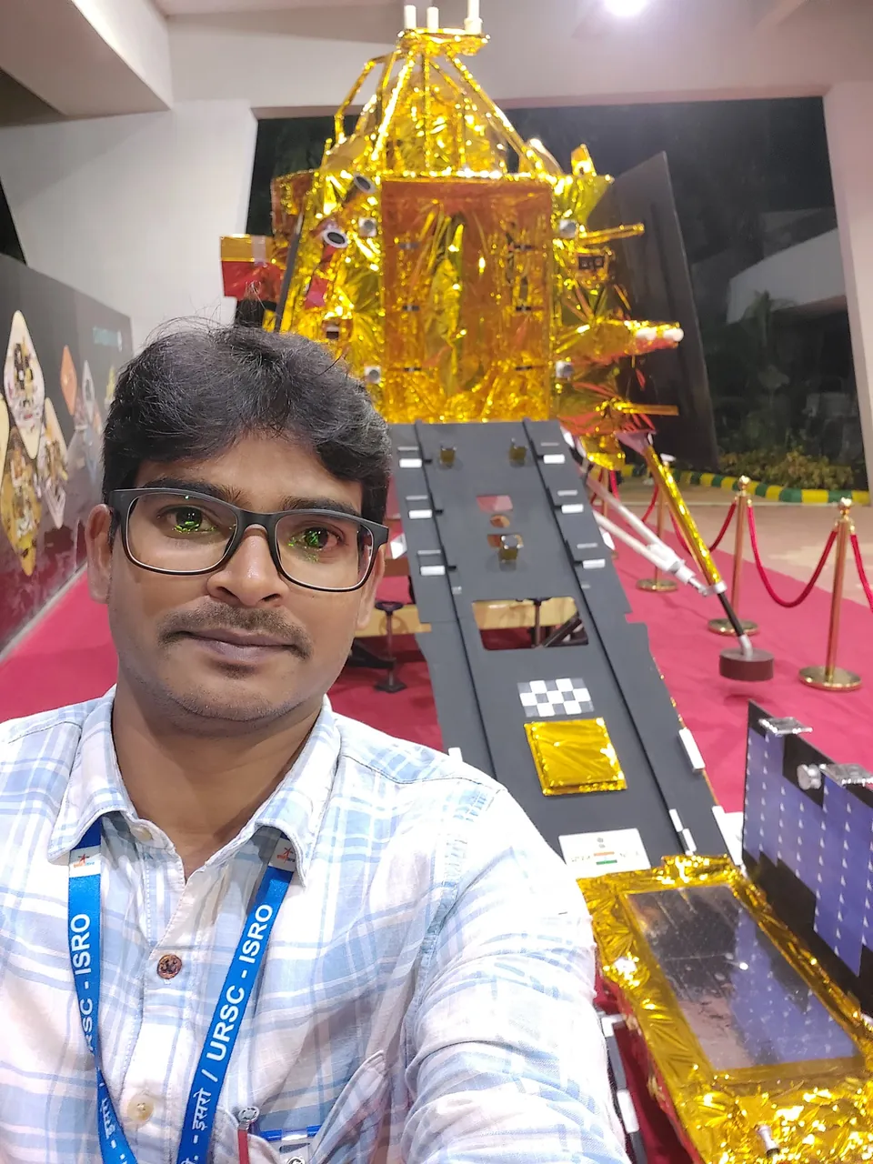 Selfie of Krishna Kummari in front of a gold-foiled lander model with a ramp leading down from it.
