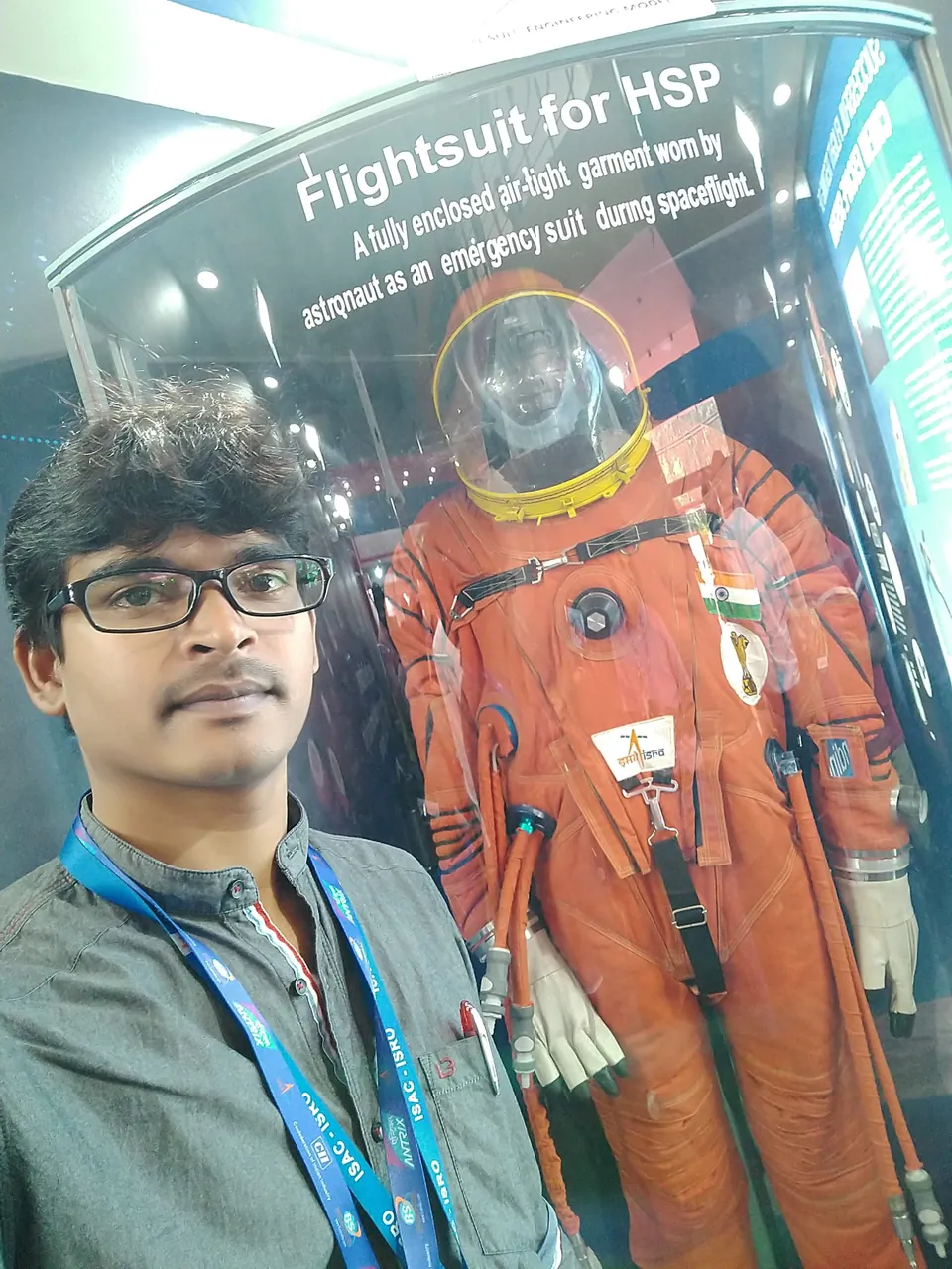 Selfie of Krishna Kummari beside a display case holding an orange flight suit labelled Flightsuit for HSP.