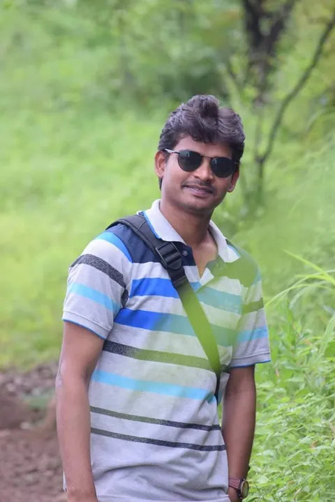 Krishna Kummari outdoors among green trees, wearing sunglasses, a striped T-shirt and a backpack.