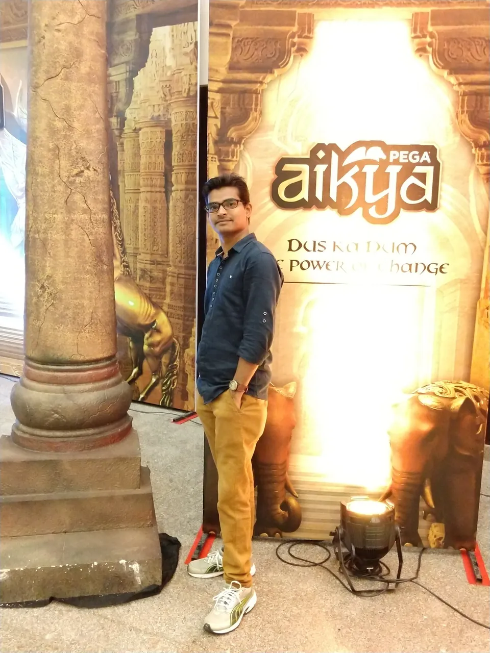 Krishna Kummari standing beside a lit stage backdrop with the words Pega Aikya.