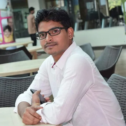 Krishna Kummari in a white shirt and glasses, seated at a table and smiling slightly.