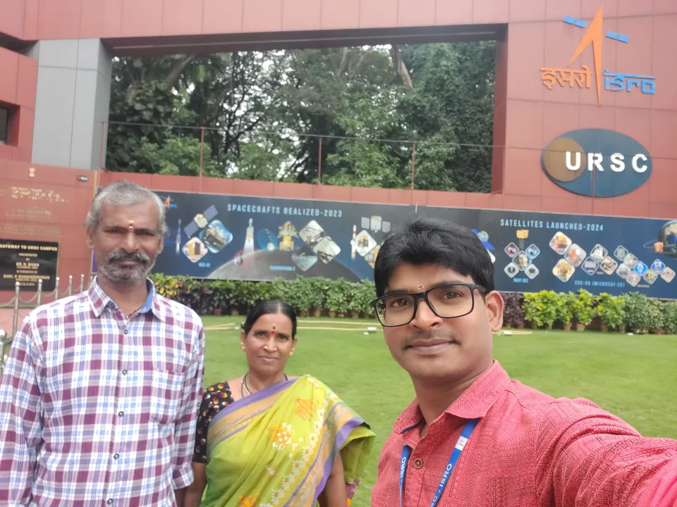 Krishna Kummari taking a selfie with his parents in front of the URSC entrance sign, a lawn and trees behind them.