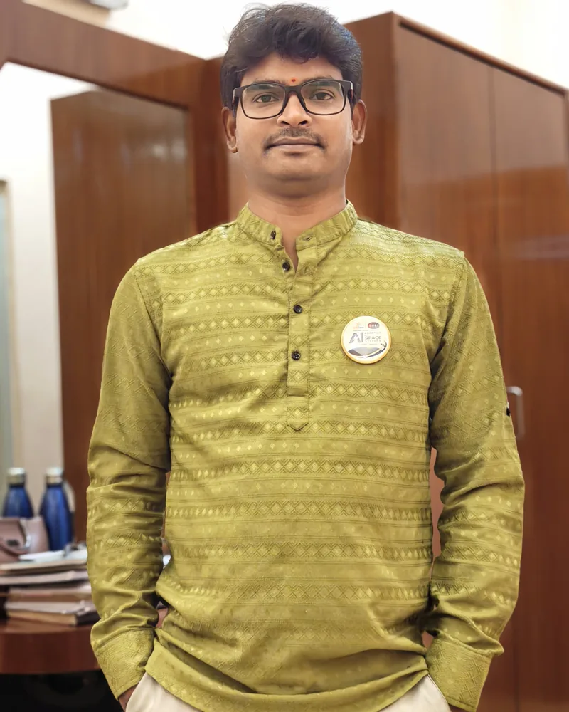 Krishna Kummari standing indoors in an olive-green patterned kurta, wearing dark-rimmed glasses.